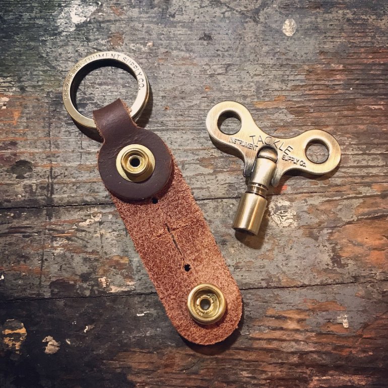 Timekeepers Drum Key from Tackle shown in antique brass unpacked at an old wooden surface with its leather sleeve next to it